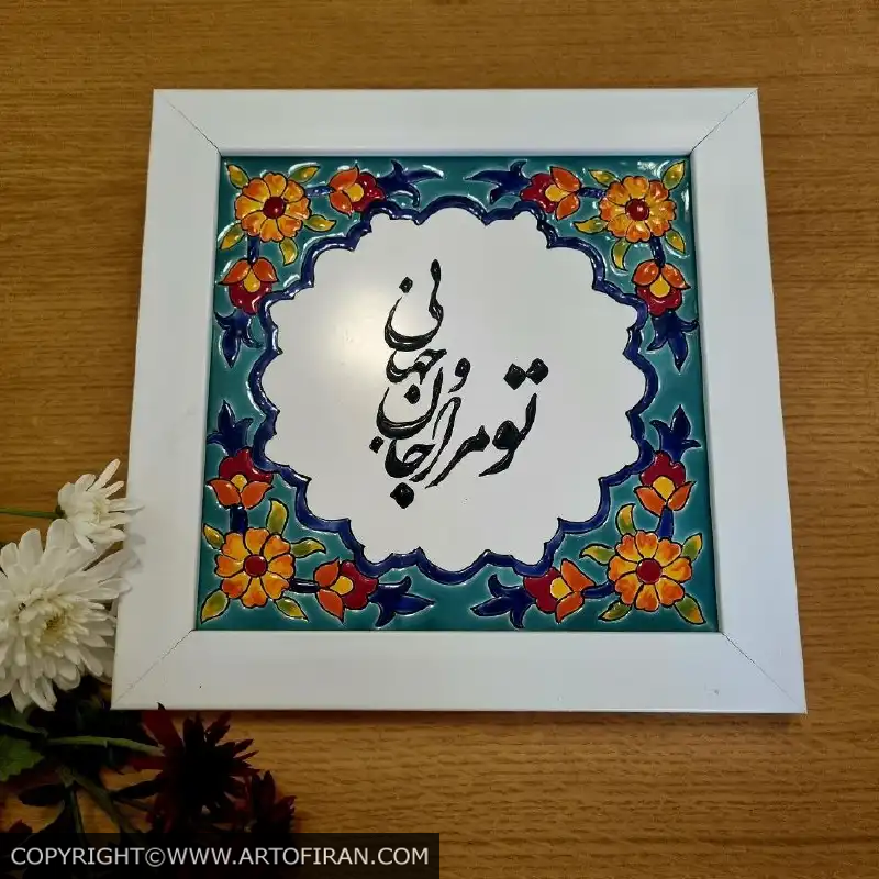 نمای کاشی هفت رنگ جام و جهان با قاب سفید روی میز چوبی، کنار گلهای سفید و قرمز